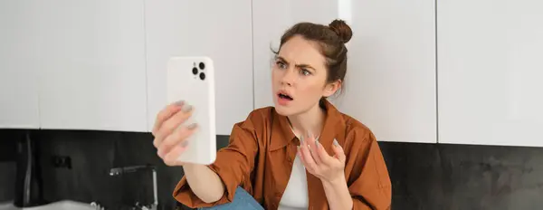 Portrait of woman with confused face, talking at smartphone, video chats, expresses frustration and disappointment.