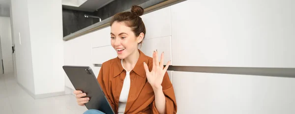 Portrait of woman saying hello, waving hand at digital tablet, connects to video chat, studying online on remote using her device to connect to the class.