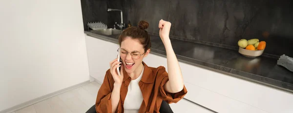 Happy woman receive positive news, job offer over phone call, celebrating, sitting at home, talking on telephone with rejoice, dancing on her seat.