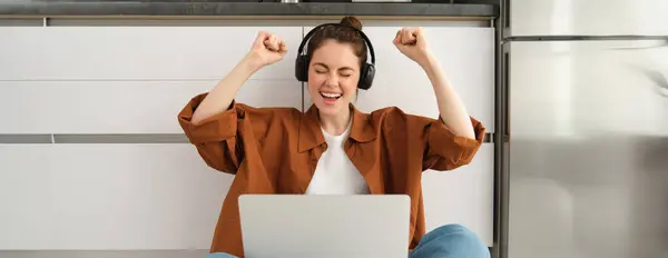 Enthusiastic young woman achieves goal, sits on floor with laptop and headphones, screams with excitement, wins in game on computer, celebrates victory, triumphs.