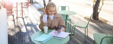 Genç güzel bir kız resmi, öğrenci çalışıyor, kafede ödev yapıyor, cep telefonu kullanıyor, akıllı telefon tutuyor, gülümsüyor..