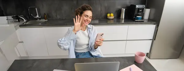 Young friendly woman, waves hand at laptop camera, video chats, holds smartphone, has conversation online.