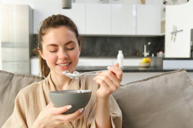 Gülümseyen, kahvaltı eden, elinde mısır gevreğiyle evde yemek yiyen genç bir kadın..