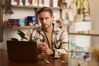 Kafe sahibi portresi, kafeteryada dizüstü bilgisayarla oturan genç bir adam, veri projesi üzerinde çalışıyor, ekrana odaklı bakıyor. Yaşam tarzı ve iş konsepti