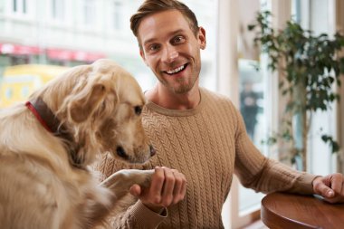 Gülümseyen genç adam köpeğinin patisini elinde tutuyor ve mutlu görünüyor. Hayvan dostu kafe ve serbest çalışma alanı kavramı.