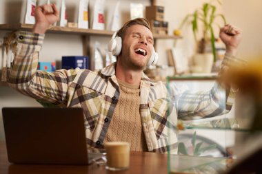 Kafede tezgahın arkasında oturan, laptopta çalışan, kahve içen, kablosuz kulaklıkla müzik dinleyen, yüksek notalar çalan heyecanlı, mutlu genç adamın portresi..