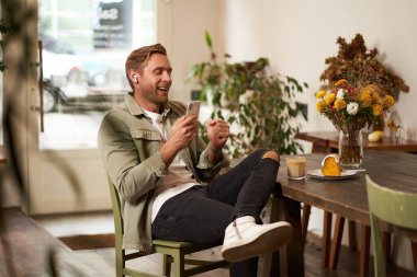 Kafede cep telefonuyla oturan, kablosuz kulaklık takan kahve dükkanında dinlenirken müzik ya da podcast dinleyen mutlu genç adamın portresi..