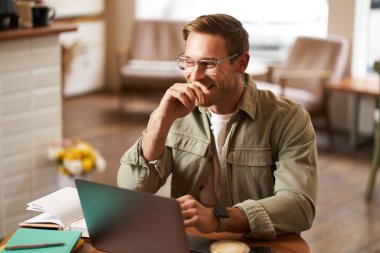 Genç dijital göçebe imajı, gözlüklü adam kafede oturuyor, kahvecide çalışıyor, iş yerinde dizüstü bilgisayar kullanıyor, gözlük takıyor, içkisini içiyor..
