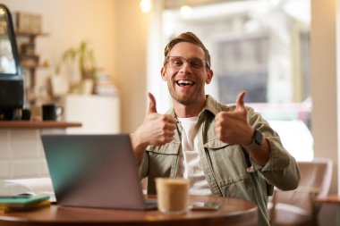 Yakışıklı gülümseyen bir adamın portresi, baş parmağını kaldırır, bir fincan kahve ve dizüstü bilgisayarla kafede oturur, güzel şeyleri onaylar, birlikte çalışma alanı veya internet sitesi tavsiye eder.