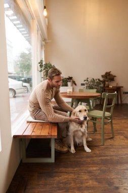 Kafede köpeğiyle oturan yakışıklı adam. Bir adam kahve içiyor ve golden retriever 'ı okşuyor. Hayvan dostu kahveler..