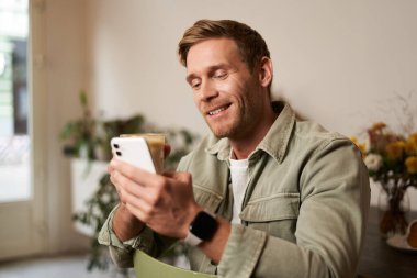 Kafedeki yakışıklı genç adamın görüntüsü, cep telefonuna bakıyor, akıllı telefon uygulaması kullanıyor, kahve içiyor..