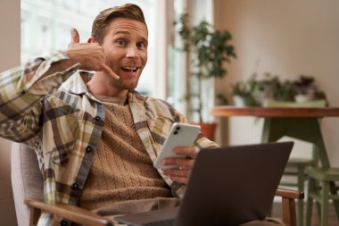 Komik, yakışıklı bir kafe ziyaretçisinin portresi, dizüstü bilgisayarı ve akıllı telefonu olan genç adam, cep telefonu işaretini gösteren ve kameraya gülümseyen, onu aramak isteyen, telefonla soran..