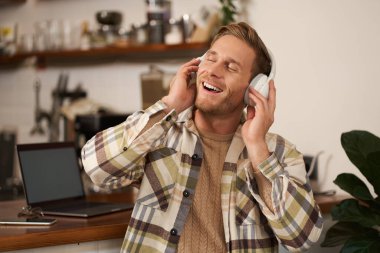 Yakışıklı, gülümseyen genç bir adam, dijital göçebe, açık alanda birlikte çalışıyor, kafeteryada çalışıyor, kablosuz kulaklıkla müzik dinliyor ve şarkı söylüyor..