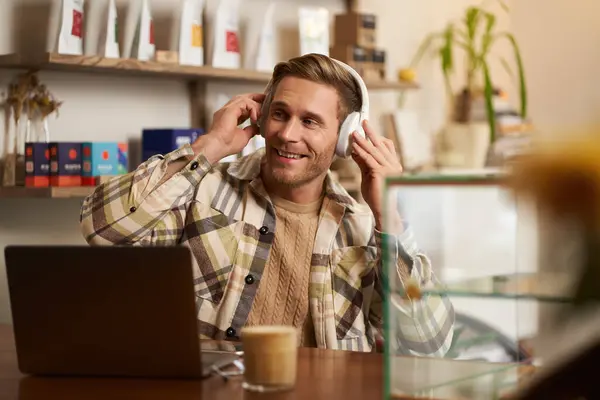 stock image Lifestyle portrait of happy, excited young man, sitting in coffee shop with laptop and headphones, dancing on his chair and enjoying great quality sound, streaming favourite songs.