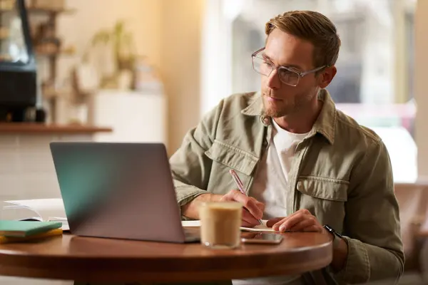 Portrait of handsome young guy in glasses, man studying, taking notes, looking at laptop screen during online lesson, attends a meeting and writing down information, sitting in cafe.