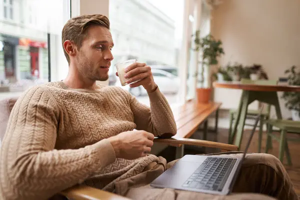 Working people and public space concept. Young handsome man, ux ui designer sitting with laptop in cafe, drinking cappuccino, looking at screen with serious face.