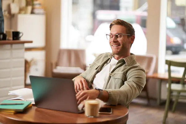 Portrait of man working in cafe on laptop, looking satisfied and pleased with himself, wearing glasses for computer screens, sits back on chair in coffee shop.