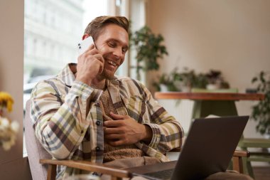 Genç profesyonel, kafeterya ziyaretçisi laptopla oturmuş müşteriyle cep telefonuyla konuşuyor. İnternetten bir şey alan, bir arkadaşını arayan bir adam..