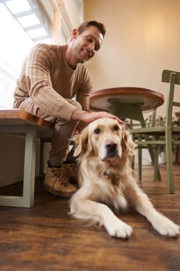 Köpeğiyle gülümseyen yakışıklı adamın dikey portresi şehirdeki hayvan dostu kafede kahve içen Golden Retriever 'a dokunuyor..