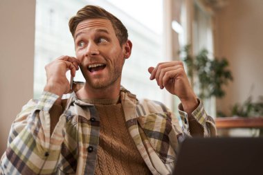 Serbest çalışma, dijital göçebe ve çalışma alanı kavramı. Cep telefonlu yakışıklı bir gencin portresi, laptopuyla kafede oturuyor, proje üzerinde çalışıyor, uzaktan çalışıyor, pencereden dışarı bakıyor..