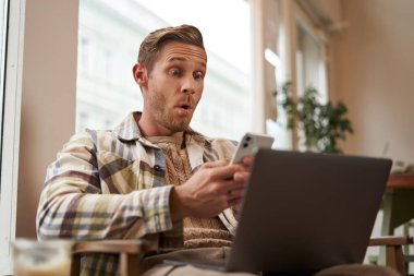 Kafedeki adamın görüntüsü, kafeterya ziyaretçisinin dizüstü bilgisayarı ve akıllı telefonuyla koltukta oturması, cep telefonu ekranına şaşırmış ve heyecanlı görünmesi, büyük haberlere hayran kalması, uygulamanın tanıtım teklifi..