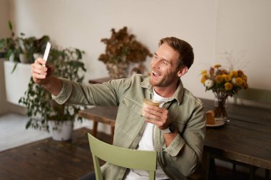 Yakışıklı genç adamın portresi, kafede selfie çekmesi, bir fincan kahve ile poz vermesi, akıllı telefon uygulaması kullanarak görüntülü konuşması, bir arkadaşla konuşması. Yaşam tarzı ve eğlence konsepti