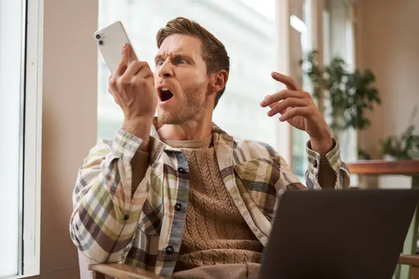 Portrait of an angry man shouting at his mobile phone. Frustrated cafe visitor arguing with someone on call, sitting with laptop near window, screaming at telephone.