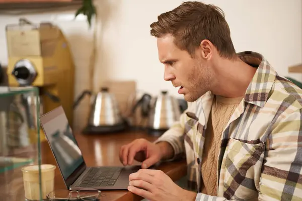 Portrait of man looks frustrated at laptop screen, staring at monitor with shocked face expression, reading message with frowned face, sitting in cafe, working remotely from coffee shop.