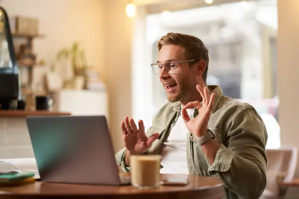 Portrait of young man, teacher, online tutor in glasses, showing okay, ok hand sign, giving lessons remotely from cafe, sitting with laptop, talking to someone on video chat.