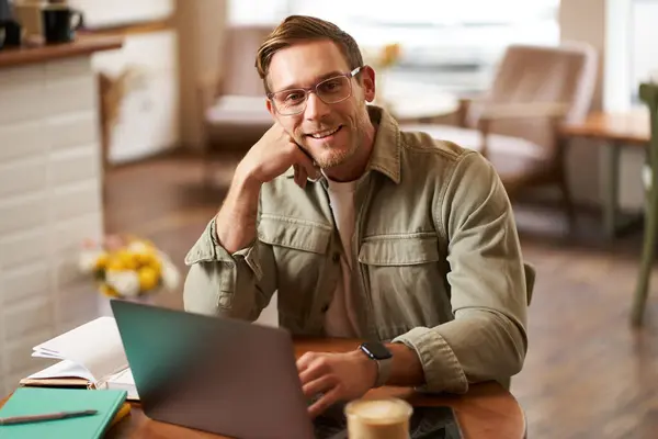 Remote work and freelance concept. Handsome man in glasses sits with notebook and laptop, studying online, working remotely from coffee shop, sitting in cafe with computer.