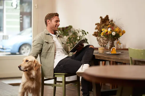 Portrait of handsome young man, cafe visitor, sitting with his dog, petting golden retriever and reading news on digital tablet, concept of lifestyle and pet-friendly public places.