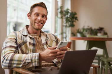 Genç, yakışıklı bir adamın portresi, kafeterya ziyaretçisi, dizüstü bilgisayarlı ve akıllı telefonlu, kameraya gülümseyen, uzaktan çalışan serbest çalışan biri..