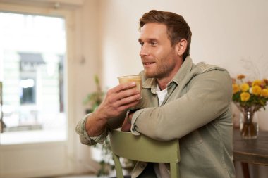 Yakışıklı bir adamın yakın portresi, sandalyede oturuyor, elinde bir bardak kahve, cappuccino içiyor ve gülümsüyor. Yaşam tarzı ve eğlence anlayışı.