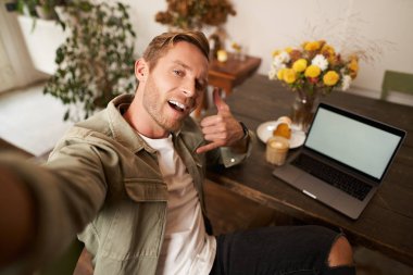 Yakışıklı adam cep telefonunda dizüstü bilgisayarla selfie çekiyor, çağrı el işareti yapıyor, kameraya gülümsüyor, sosyal medya uygulamasına fotoğraf yüklüyor, kafede oturuyor..