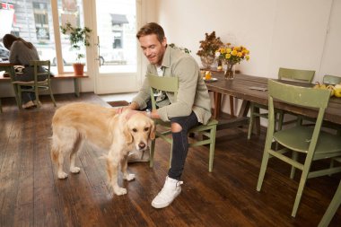 Güzel köpeğiyle evcil hayvan dostu kafede oturan yakışıklı bir adamın hayat tarzı portresi Golden Retriever 'ı okşuyor ve bir emir bekliyor..