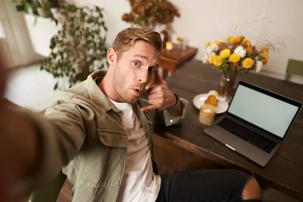 Handsome stylish young man takes selfie in cafe while working or studying remotely with laptop, shows call phone hand sign and smiling at mobile phone camera.