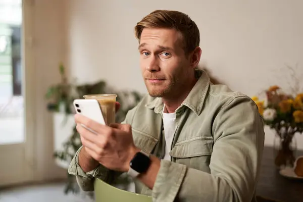 Portrait of young man in cafe, holding cup of coffee and mobile phone, looking at camera with skeptical face and a smirk.