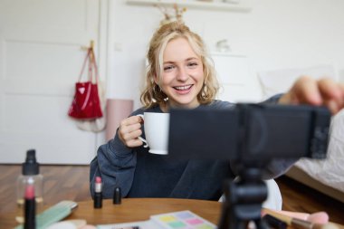 Şirin gülümseyen kadın portresi, sarışın kız çay içiyor ve sıradan bir video blogu kaydediyor, vlogger kameralı ve dengeleyicili bir odada oturuyor, elinde bir fincan kahve tutuyor ve konuşuyor..