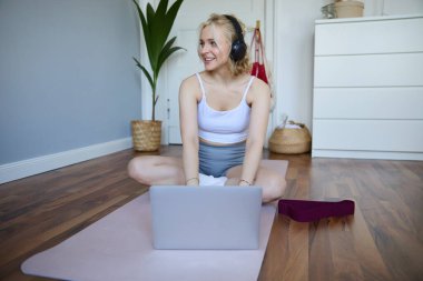 Evde gülümseyen sporcu kadının portresi, kauçuk yoga minderinde egzersiz yapmak, dizüstü bilgisayar ve kablosuz kulaklık kullanmak,.