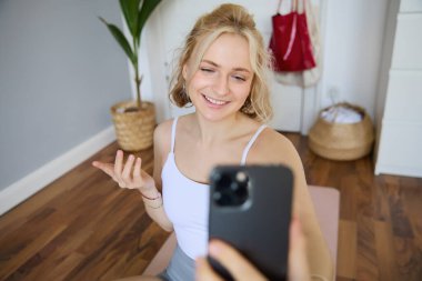 Güzel genç fitness eğitmeninin portresi, ev antrenmanı, cep telefonuyla selfie çekmek, yatak odasında yoga lastik minderinin üzerinde otururken, aktif giysi giymek..