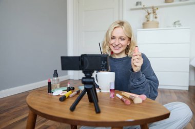 Güzel, gülümseyen sarışın kadının portresi, sosyal medya için makyaj dersinin videosunu çeken kız, odasında yerde oturan vlogger, içerik yaratmak için dengeleyici kullanıyor, rimelini gözden geçiriyor..