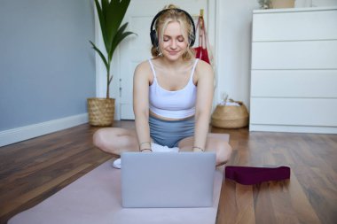 Spor yaparken, spor yaparken, dizüstü bilgisayar kullanırken, yoga minderinde otururken çevrimiçi video talimatlarını izleyen genç kadının portresi..