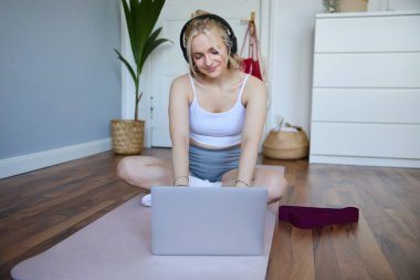 Evde gülümseyen sporcu kadının portresi, kauçuk yoga minderinde egzersiz yapmak, dizüstü bilgisayar ve kablosuz kulaklık kullanmak,.