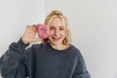 Yüzünün yanında pembe donut gösteren, gülümseyen ve mutlu görünen genç sarışın bir kadının yakın portresi. Halk konsepti