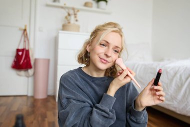 Kadın güzellik blogcusu, kendini bir odada otururken ve makyaj ürünlerini değerlendirirken videosunu kaydediyor, makyaj yapıyor, ruj sürüyor ve makyaj fırçasını elinde tutuyor, içerik yaratmak için profesyonel kamera kullanıyor..