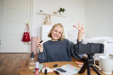 Genç sarışın kadının portresi, genç kız sosyal medya hesabı için video kaydediyor, kamerada makyaj gösteriyor, online takipçilere ruj öneriyor, odasında içerik yaratıyor..