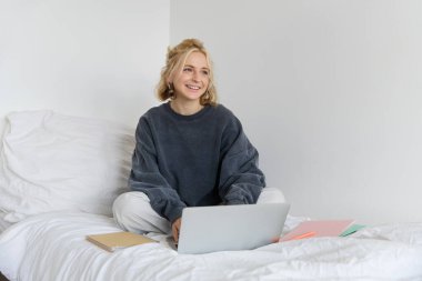 Mutlu genç bir kadın resmi, evden öğrenim görüyor, laptopuyla online kursa bağlanıyor, not defterleriyle yatakta oturuyor, gülümsüyor ve mutlu görünüyor..
