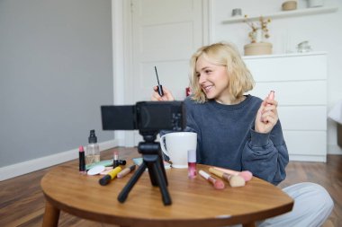 Odasında gülümseyen genç bir kadının portresi, kamerada video kaydı, sosyal medya için yaşam tarzı vlog, rimel tutmak, makyaj ürünlerini incelemek, kozmetik ürünlerin nasıl kullanılacağını göstermek..