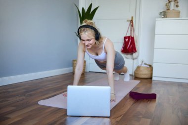 Spor yapan genç bir kadının portresi, kablosuz kulaklıkla dizüstü bilgisayardaki fitness videolarına bakıyor, yoga minderinde tahtada duruyor,.