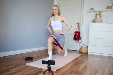 Spor eğitmeninin portresi, kamera önünde egzersiz yapan genç vlogger kadın egzersiz videosu çeken, direnç bandını kullanan ve boş bir odada mekik çeken kadın..